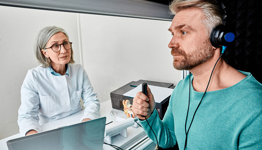 A Male Patient Sitting With an Audiologist Wearing Headphones & Holding a Response Button During a Hearing Test Types of Hearing Test