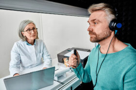 A Male Patient Sitting With an Audiologist Wearing Headphones & Holding a Response Button During a Hearing Test Types of Hearing Test