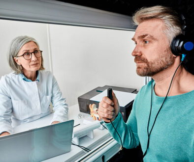 A Male Patient Sitting With an Audiologist Wearing Headphones & Holding a Response Button During a Hearing Test Types of Hearing Test