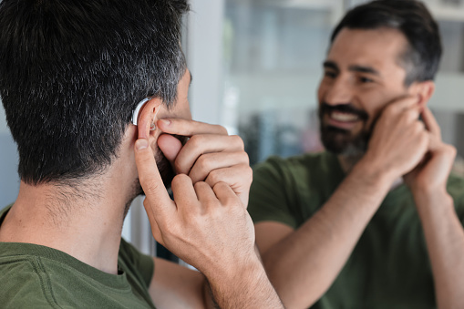Handsome man with BTE hearing aid looks at herself in mirror and tries on hearing device, close-up. Hearing loss treatment