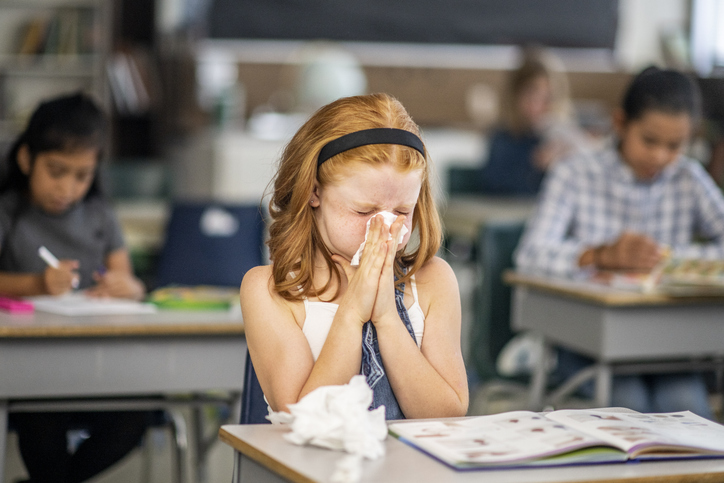 Focus is on a female child in the classroom. She has a tissue to her nose and is sneezing. Other children are also in the background and seated, but out of focus.
