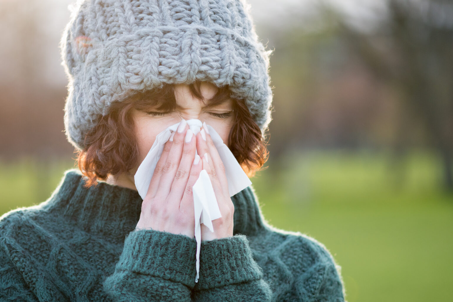 Woman wearing a sweater and knit beanie blowing her nose on a fall day.